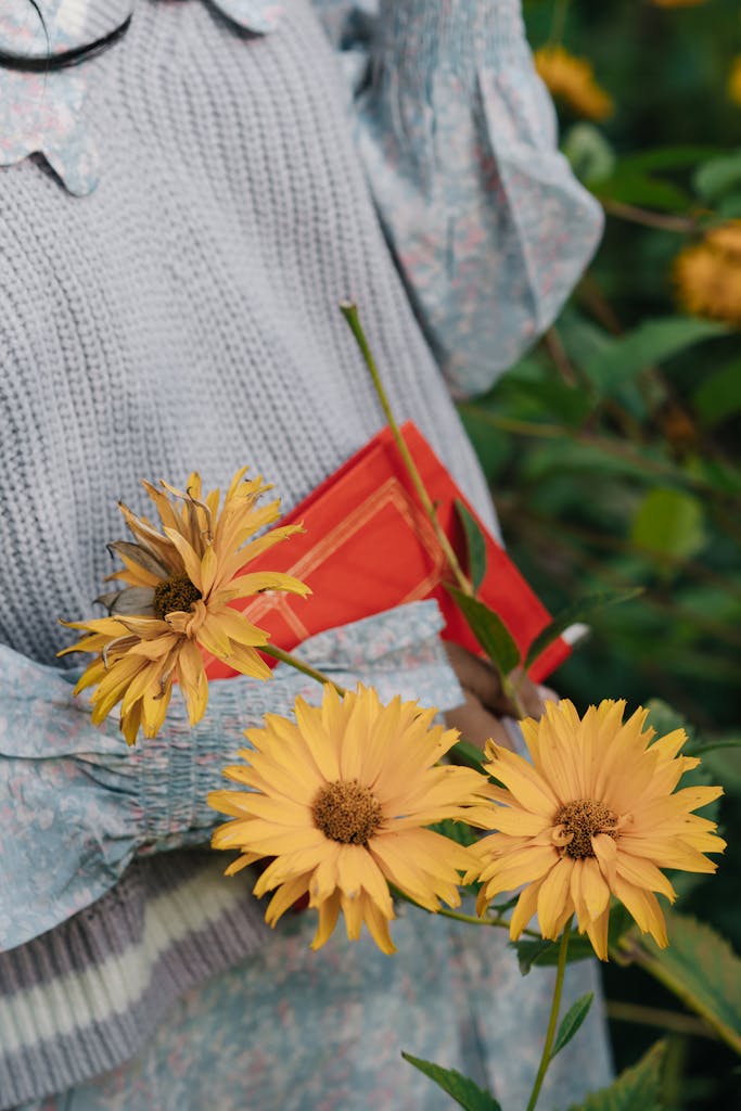 A woman in a floral dress holds vibrant yellow chrysanthemums outdoors.
